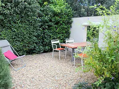 Terrasse de jardin en gravier avec table, chaises blanches et chaise longue au coussin rose devant une haie verdoyante.