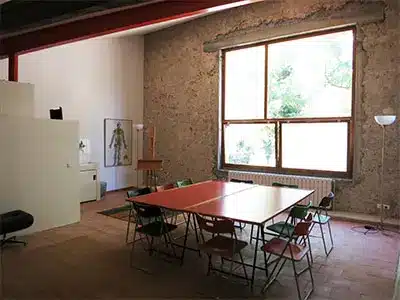 Atelier d'artiste avec grande table rouge, chaises pliantes, mur en pierre et large fenêtre lumineuse.