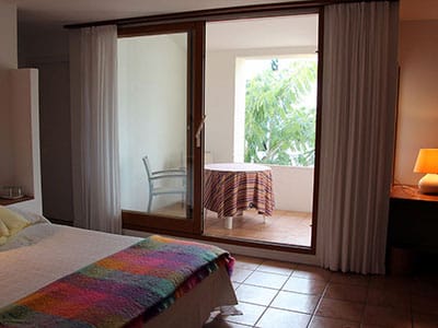 Chambre lumineuse avec lit coloré ouvrant sur un balcon aménagé avec table et vue sur jardin tropical.