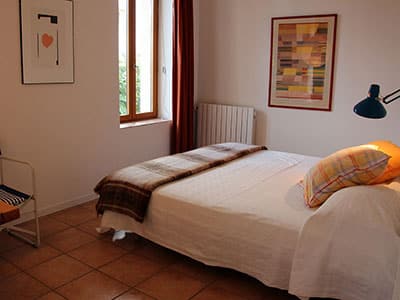 Chambre lumineuse : lit double avec jeté à carreaux, oreillers colorés, tableaux décoratifs et fenêtre aux rideaux rouges.