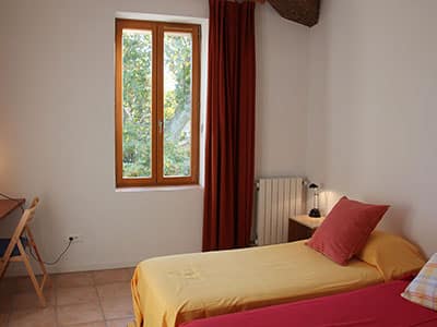Chambre avec lits simples colorés, rideau rouge, fenêtre en bois et chaise pliante sur sol en terre cuite.