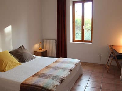 Chambre lumineuse avec lit double, couverture à carreaux, fenêtre en bois et sol en carrelage de terre cuite.