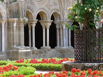 Jardin du cloître avec arches romanes et fleurs Cloître aux arches romanes et colonnes sculptées surplombant un jardin de fleurs rouges et un rosier en fleurs.