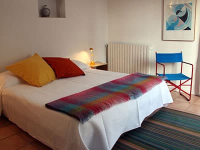 Chambre lumineuse avec lit blanc, oreillers jaune et rouge, plaid coloré et fauteuil bleu sur tapis rayé.