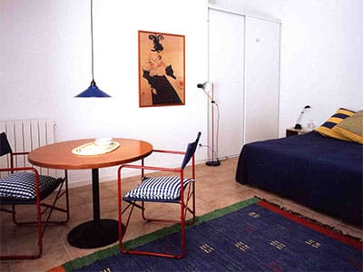 Studio lumineux avec table ronde en bois, chaises rouges, lit bleu et grand tapis décoratif.