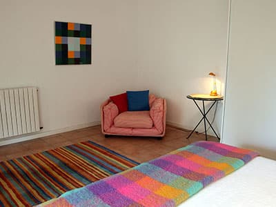 Coin lecture avec fauteuil rose, tapis multicolore, tableau géométrique et couverture à carreaux colorée.