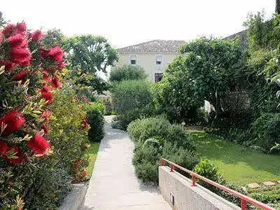 Jardin luxuriant avec rince-bouteille rouge, chemin pavé menant vers une maison méditerranéenne sous un ciel clair.