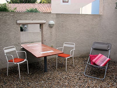 Table en terre cuite, chaises blanches et transat sur une terrasse en gravier avec mobilier de jardin moderne.