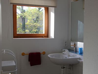 Salle de bain lumineuse avec lavabo blanc, fenêtre vue jardin et serviette rouge sur un porte-serviette jaune.