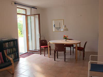 Salle à manger lumineuse avec table en bois, chaises bordeaux, bibliothèque et porte ouverte sur un jardin.