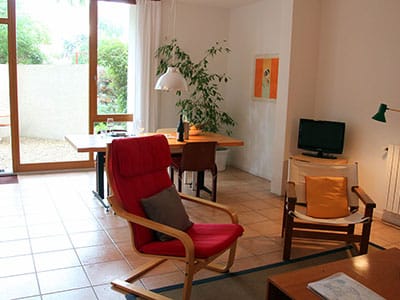 Salon lumineux avec fauteuil rouge, table à manger en bois et grande baie vitrée ouvrant sur un jardin.