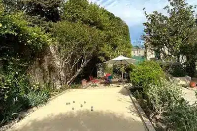 Terrain de pétanque privé au cœur d'un jardin luxuriant Terrain de pétanque ombragé dans un jardin luxuriant avec boules, parasol blanc et chaises rouges.